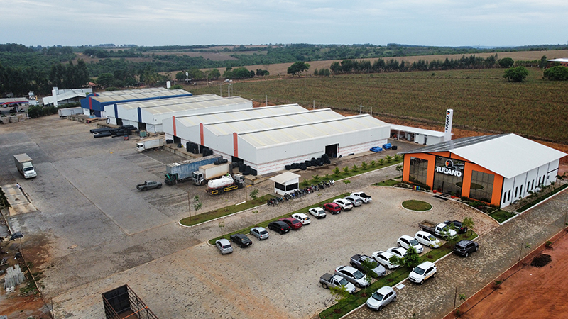 Vista aérea do complexo industrial da Tucano Tubos, exibindo galpões extensos, área de estacionamento com diversos carros e uma fachada de escritório com o logotipo da empresa.